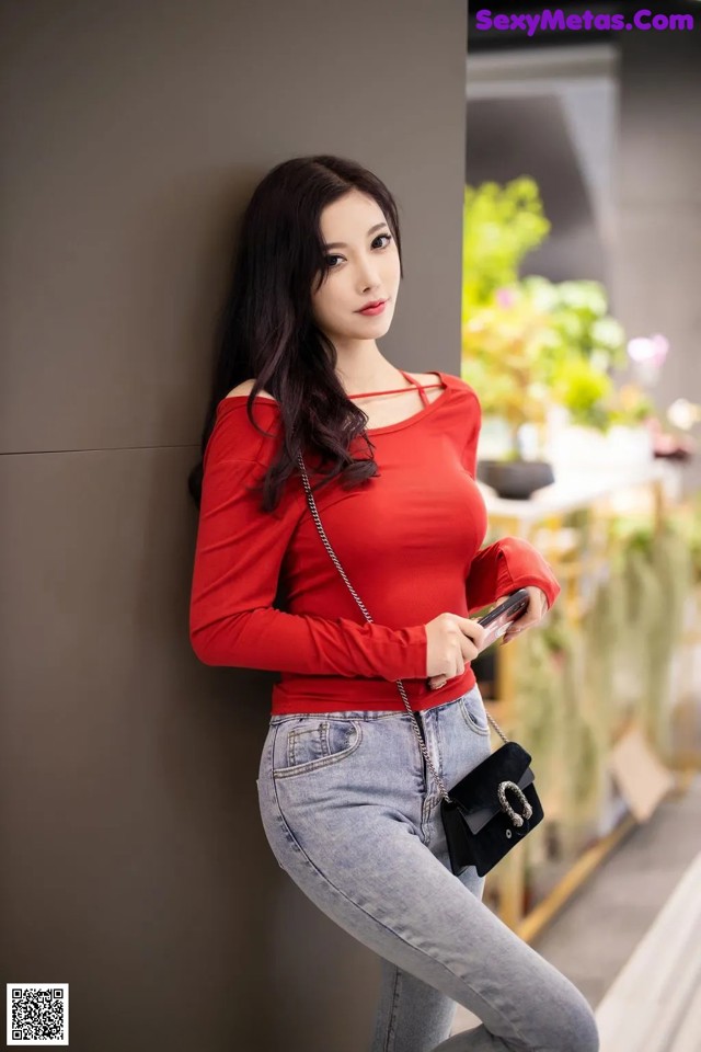 A woman in a red top and jeans leaning against a wall.