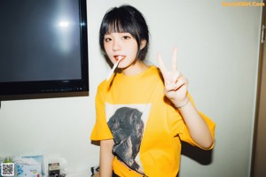 A woman standing in front of a bed wearing a yellow shirt with a picture of Paul McCartney on it.